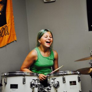 8 year old, female student with green hair in a ponytail and a green tank top is playing a white drum kit.
