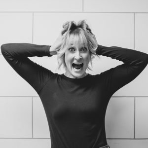 Woman with short blonde hair holding both hands to the sides of her head with surprised expression on her face.