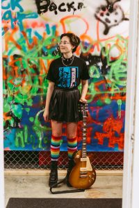 Teenager with short brown, curly hair and glasses standing with electric guitar next to him. Wearing rainbow-coloured knee high socks and black pleated leather skirt and black T-shirt.