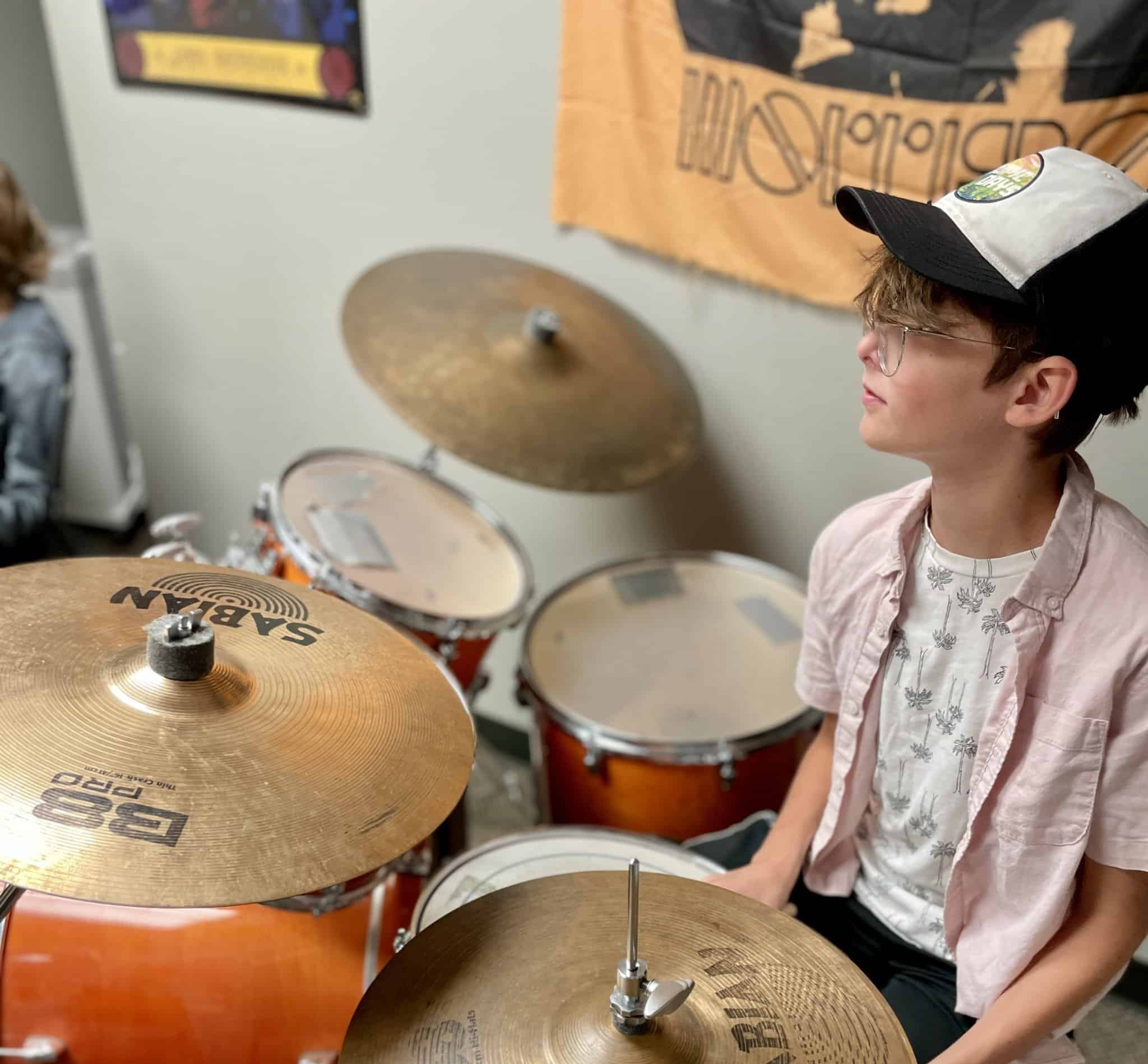 10 year-old, male student with short blonde hair is wearing a baseball cap and is sitting at orange drum kit.