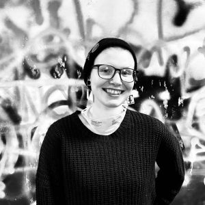 Jess Tollestrup, black and white photo. They are wearing a black tocque, glasses and black sweater. They have a mask hanging around their neck. They have a big smile on their face and square, dangling earrings.