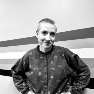 Anna Appleton. Black and white photo. Anna has a short, blonde buzz cut and has a pleasant look on her face with a smile. Behind Anna is a rainbow wall.