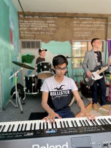 Teenage boy with glasses and black hair plays keyboard with his rock band in the back. Performance is outdoors in a back alley.