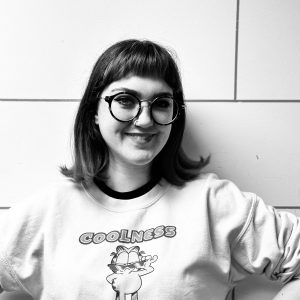 Black and white photo of a young adult with shoulder length hair and thick rimmed glasses.