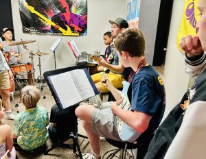 A group of nine and ten year old kids playing electric guitar, keyboards and drums under the direction of a teacher.