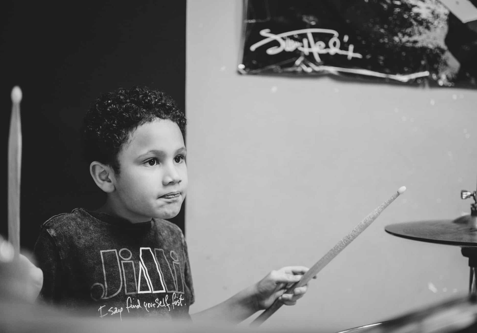Axton Aryee playing drums in Edmonton. Black and white photo og young boy with light curly dark hair playing drums. He has a focused look on this face.