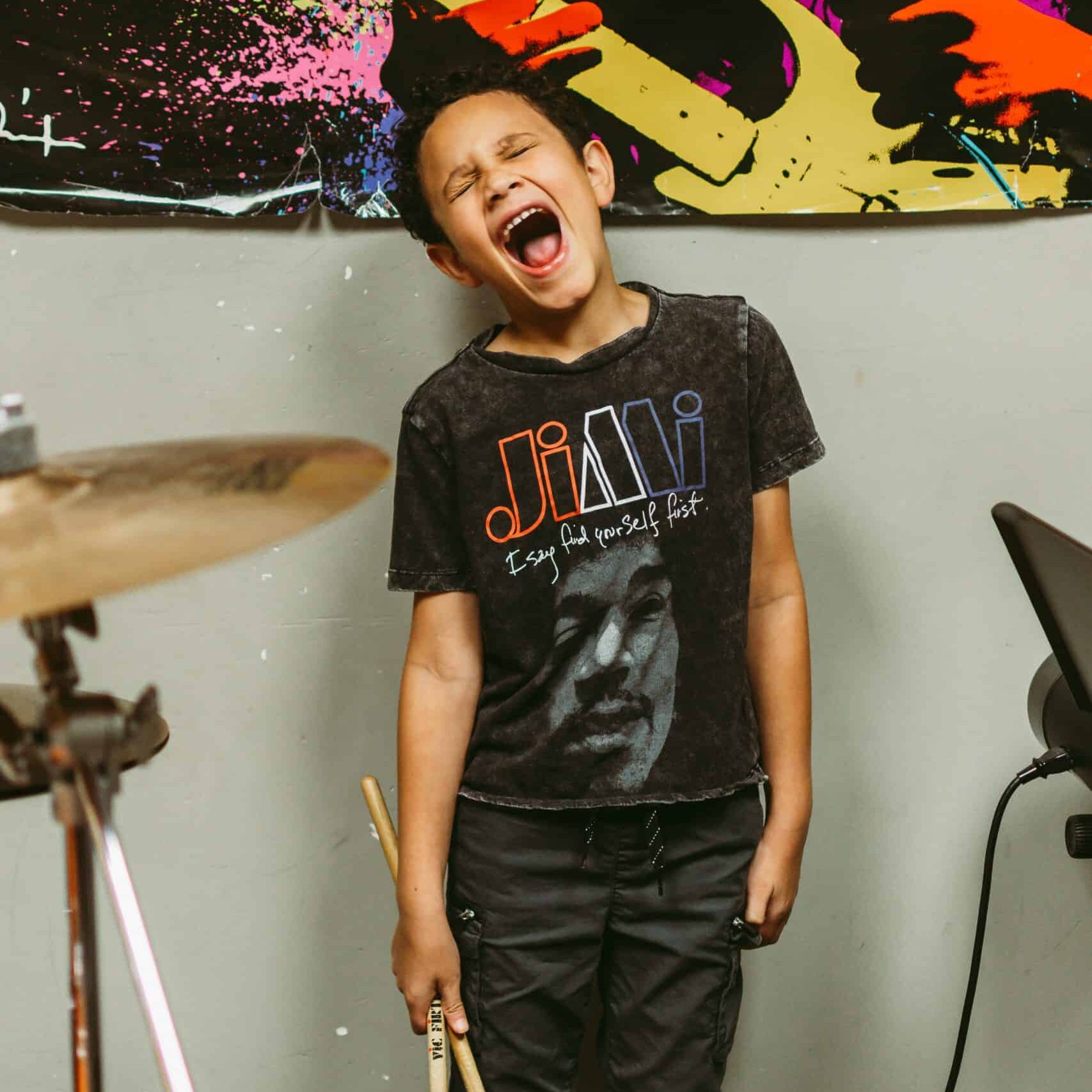 Axton Aryee // taking drum lessons at Backbeat in Edmonton Young boy with light brown skin and curly black hair is standing next to a drum kit screaming in excitment while holding a pair of drum sticks.