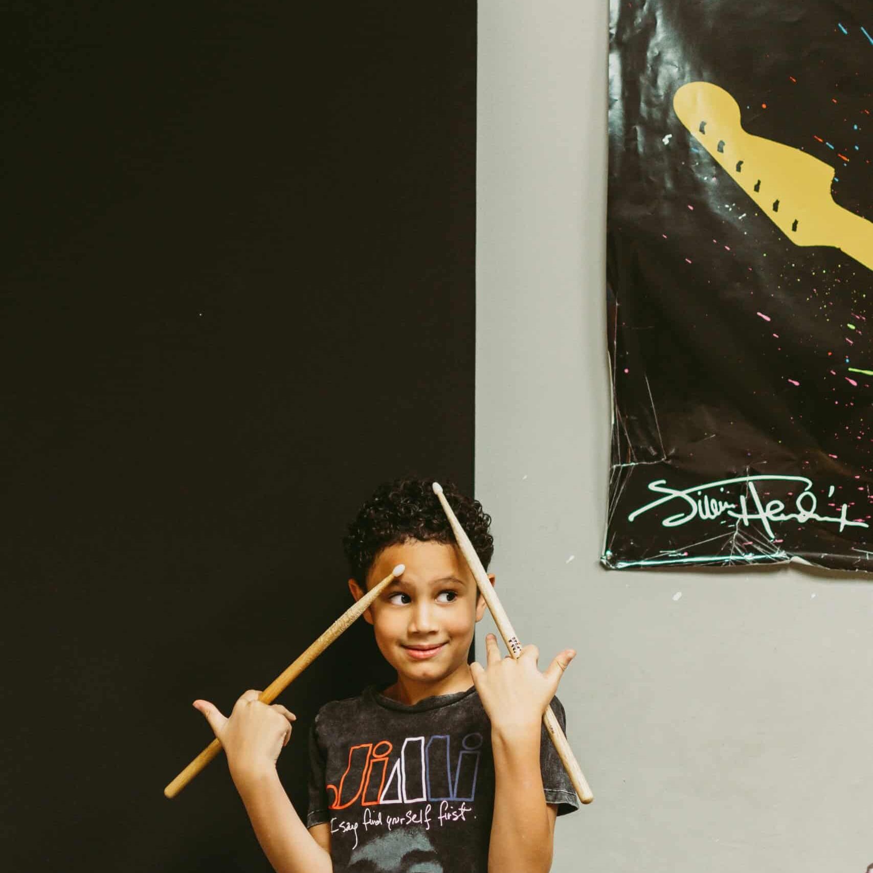 Axton Aryee is taking taking drum lessons at Backbeat in Edmonton. Young boy with light brown skin and black, curly hair is standing with a goofy look on his face next to a drum kit. He's holding two drum sticks up.