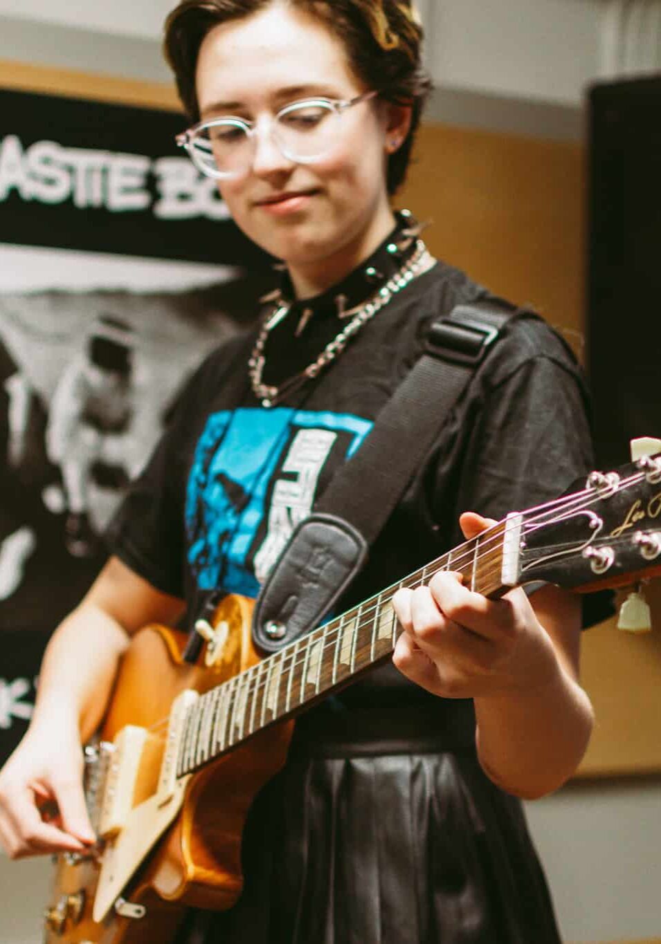 Teenager with short, curly brown hair and glasses playing orange electric guitar.
