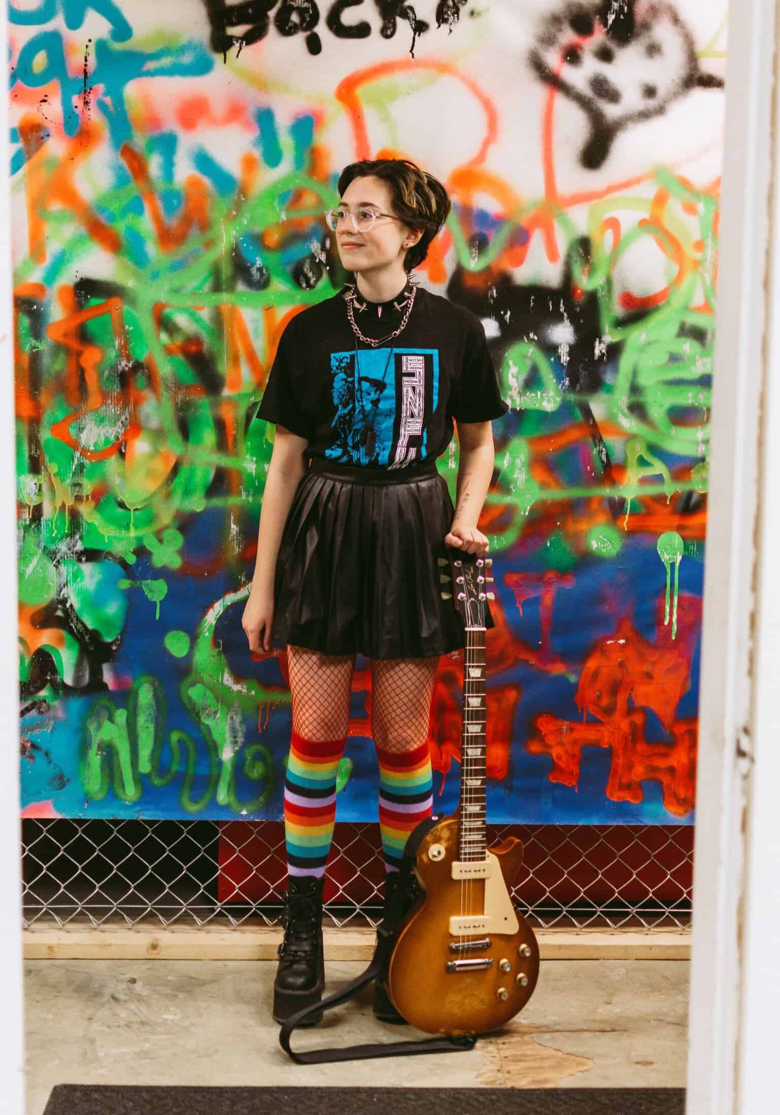Teenager with short brown, curly hair and glasses standing with electric guitar next to him. Wearing rainbow-coloured knee high socks and black pleated leather skirt and black T-shirt.
