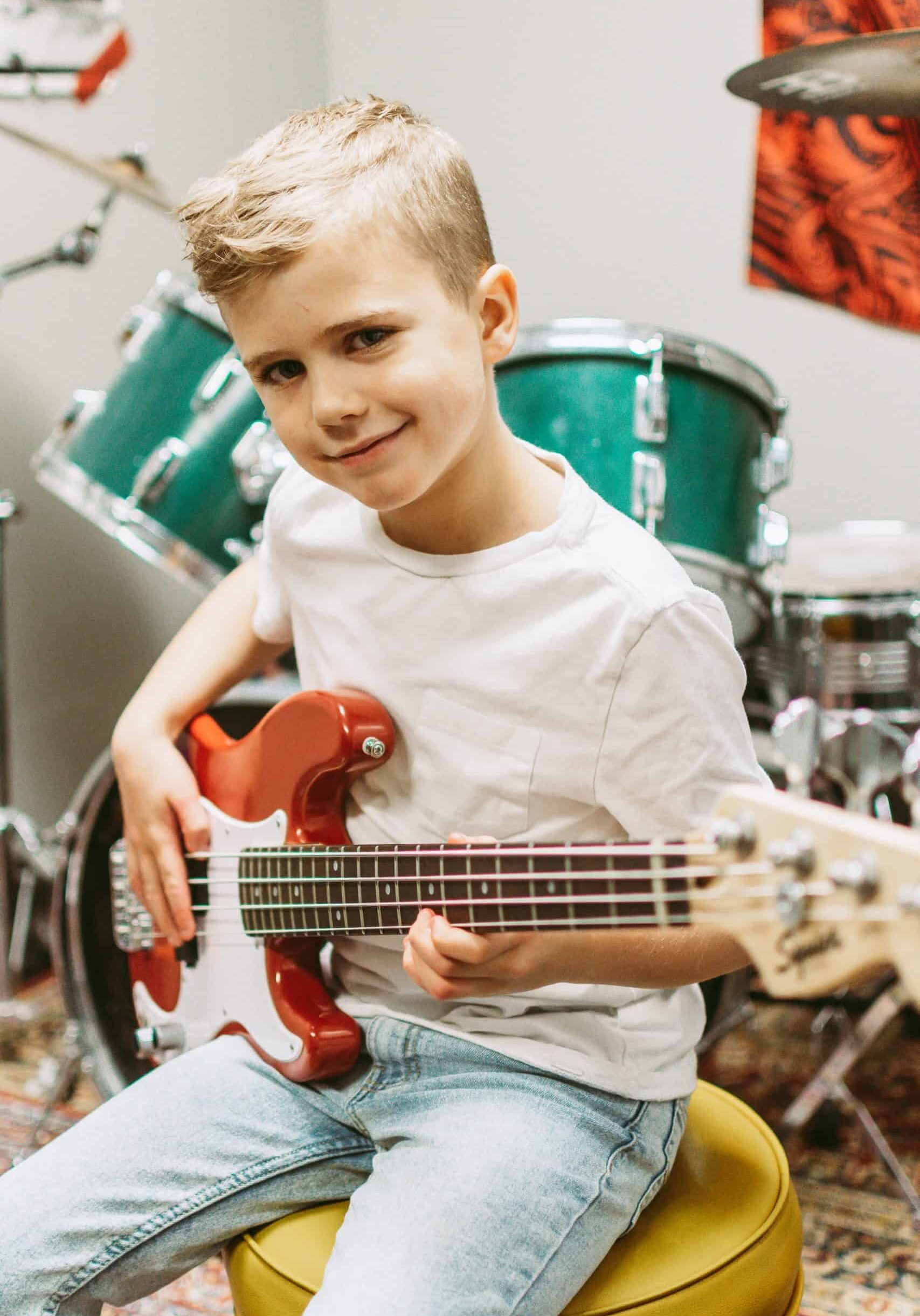 Max Leo is a bass student at Backbeat 8 year-old blonde boy sitting on a yellow stool, smiling and playing a red and white electric bass.